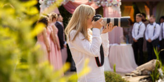 A woman wearing a white shirt holds a camera with a long lens up to her face. Out of focus in the background is a wedding party and a flowered canopy.