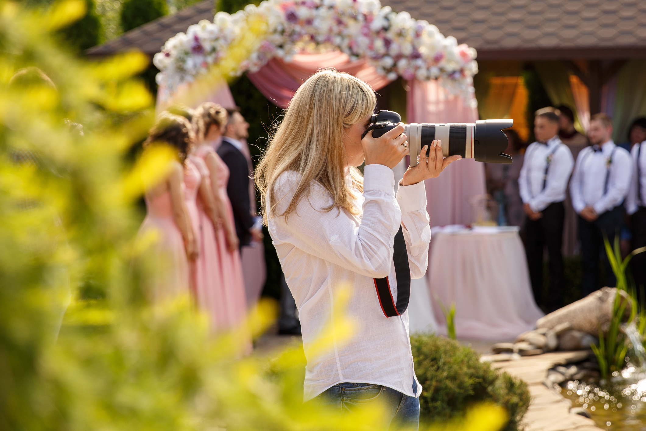 A woman wearing a white shirt holds a camera with a long lens up to her face Out of focus in the background is a wedding party and a flowered canopy
