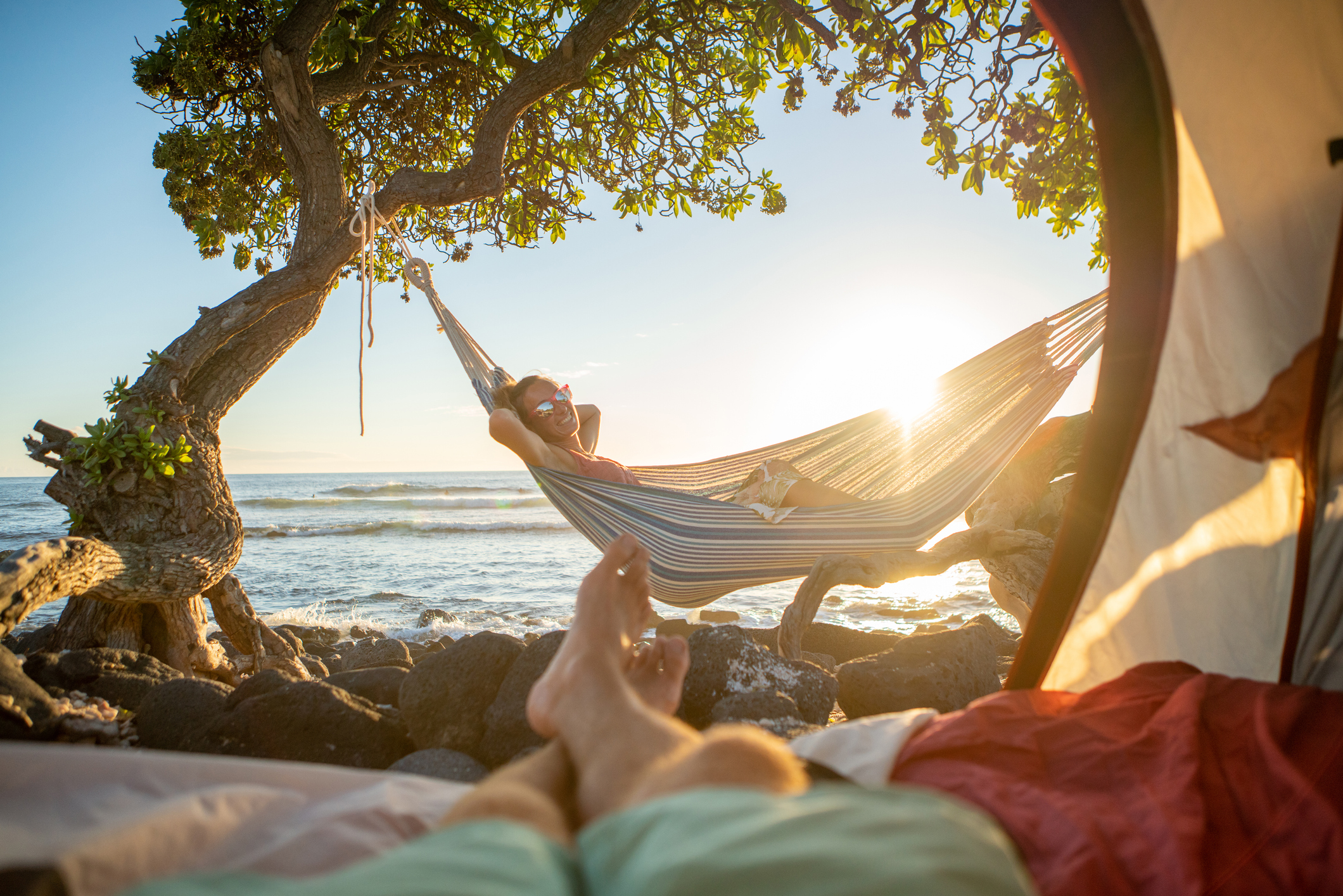This photo is taken from the perspective of someone lying on the ground inside a tent looking through the opening In the foreground you can see their legs crossed at the ankle They are looking at a woman relaxing in a hammock next to water