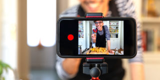 A phone held horizontally in a tripod displaying an image of a man holding several peppers. He is recording a video of himself cooking something.