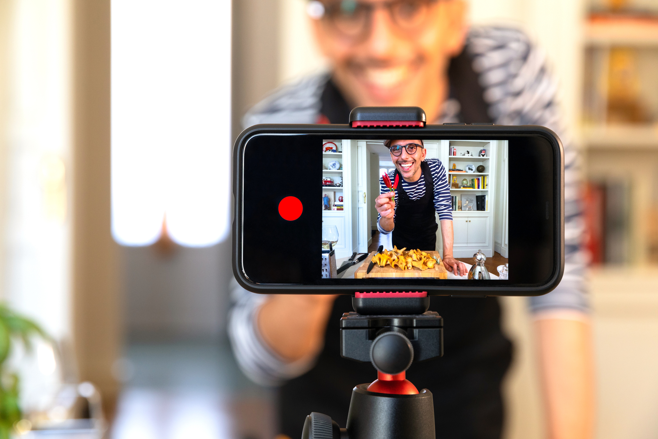 A phone held horizontally in a tripod displaying an image of a man holding several peppers He is recording a video of himself cooking something