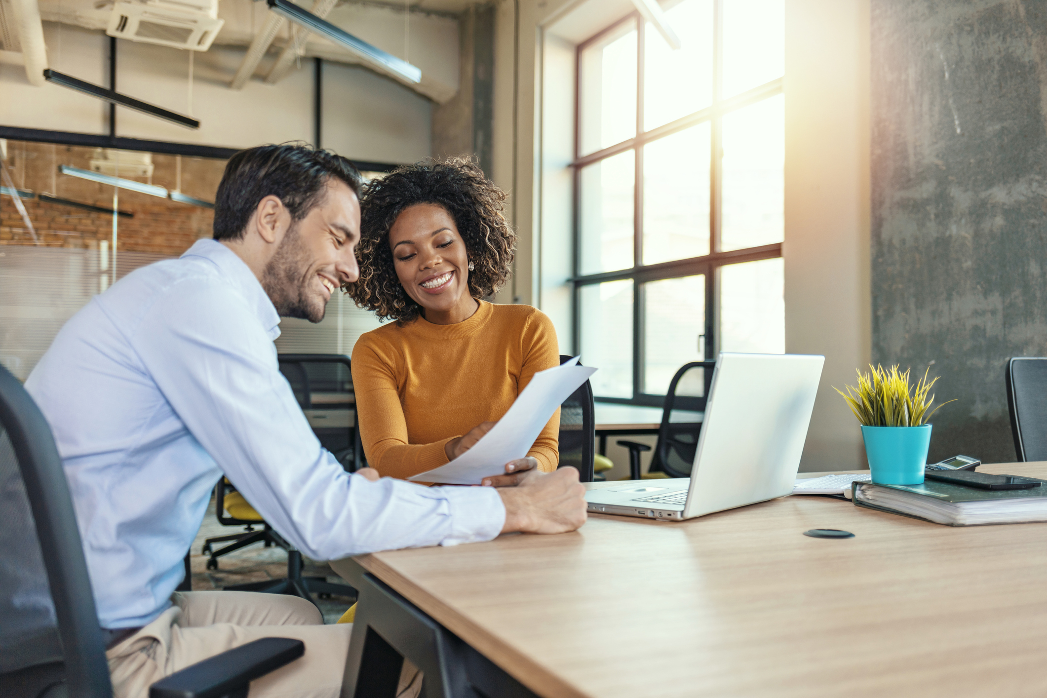 A man and woman sit at a large table reviewing paperwork in front of a computer.