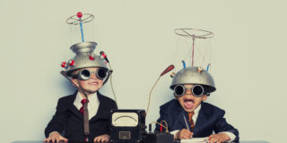 Two grinning children sit at a table wearing goggles with colanders on their heads.