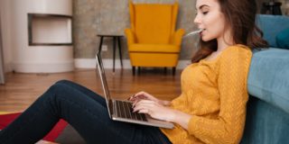 woman sits on floor leaning against sofa while performing online banking as a service on her laptop