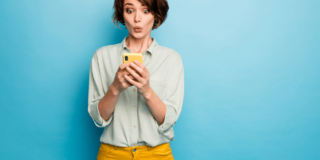 Woman in light blue top and orange pants on a blue background looking pleasantly surprised at her smartphone