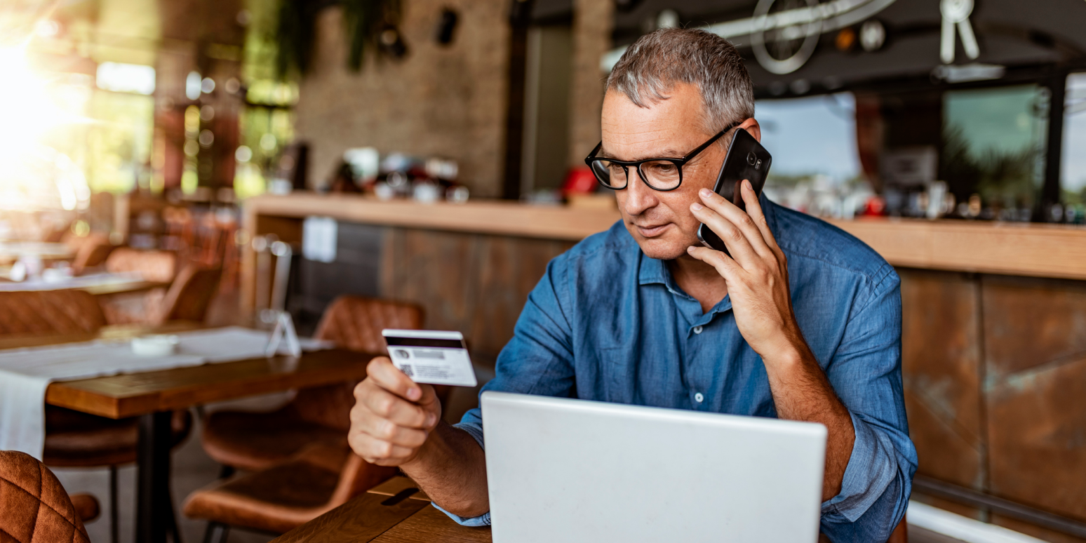 Hero image of a man at a coffee shop holding a credit card while on the phone with a computer in front of him