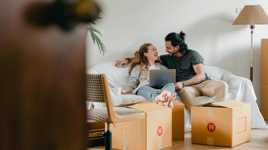 Cheerful couple in casual clothes sitting on comfortable sofa with laptop and looking at each other while moving to new apartment