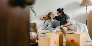 Cheerful couple in casual clothes sitting on comfortable sofa with laptop and looking at each other while moving to new apartment