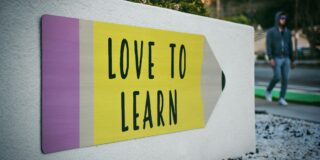 Outdoor sign on a concrete wall reading 'LOVE TO LEARN' in black letters on a yellow panel, with a blurred person in a hoodie in the background.