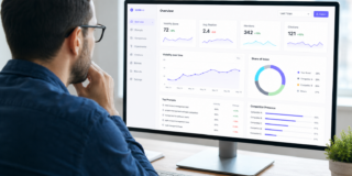 Man wearing glasses sits at a desk, reviewing a large monitor that displays an analytics dashboard with charts and metrics like visibility score and mentions.