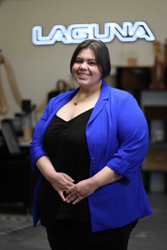 image - purshoLOGY Smiling woman in a blue blazer stands in front of a bright Laguna sign in a work setting.