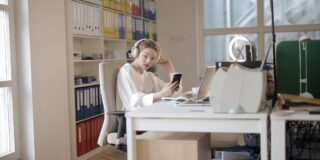 Woman with large white headphones at a desk, looking at her phone in a bright office filled with binders and a laptop.