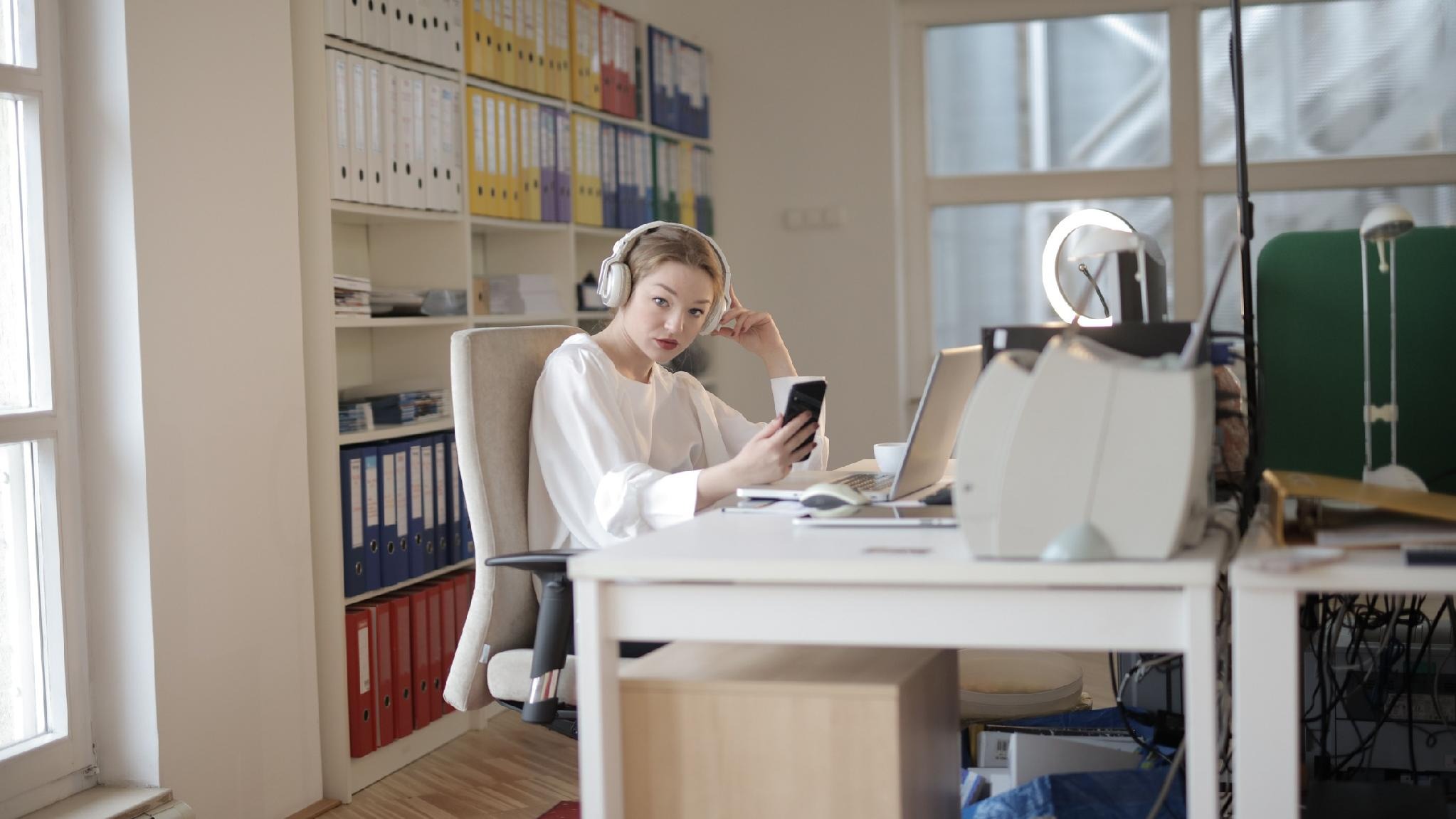 image - purshoLOGY Woman with large white headphones at a desk, looking at her phone in a bright office filled with binders and a laptop.