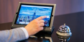 Person taps a tablet mounted in a metal frame; screen shows a city skyline and welcome text (Hello, Welcome to Upscale).