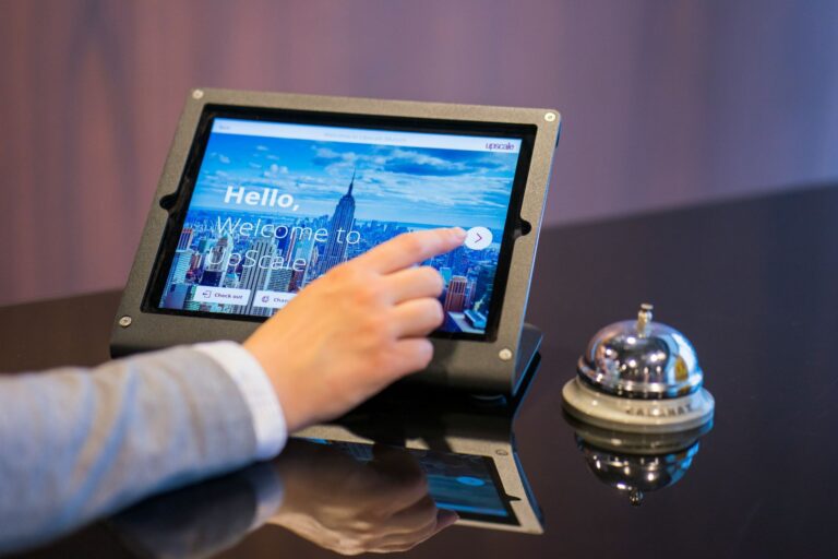Person taps a tablet mounted in a metal frame; screen shows a city skyline and welcome text (Hello, Welcome to Upscale).