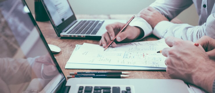Two professionals collaborate at a wooden desk with laptops and charts, one writing on a notebook with a pencil.