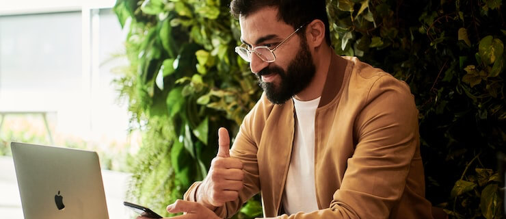 Bearded man wearing glasses giving a thumbs-up while using a laptop in a bright, plant-filled space.